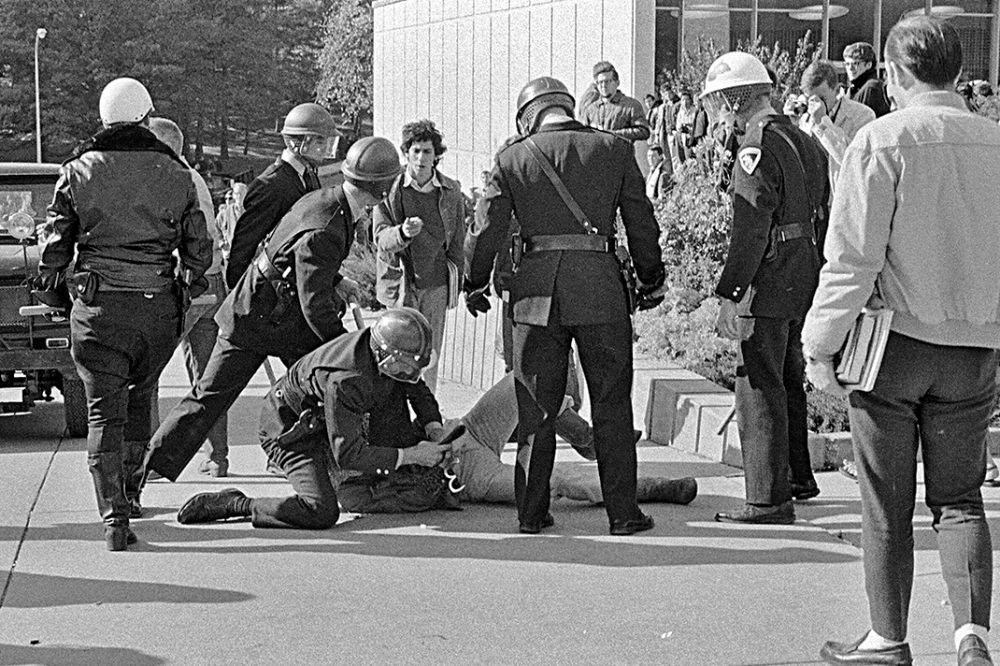 A student carrying books, perhaps on his way to or from classes, comes upon a scene of police officers attempting to handcuff a demonstrator.
