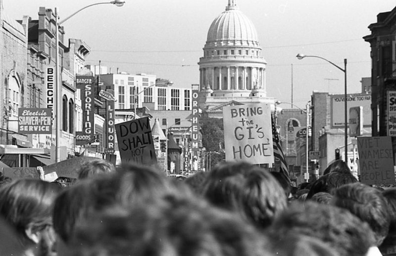 Scene from a protest looking up State Street with the State Capital in the background. Signs read "Bring the GIs Home" and "Dow Shalt Not Kill"