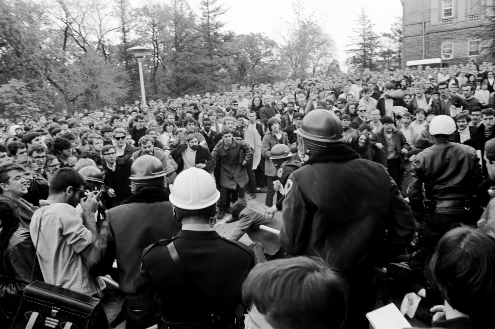 A police officer uses his billy club to strike the back of a protester as members of the crowd look on in disbelief and anger.